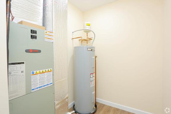 A grey water heater with a yellow warning label is in a room with a grey fridge.