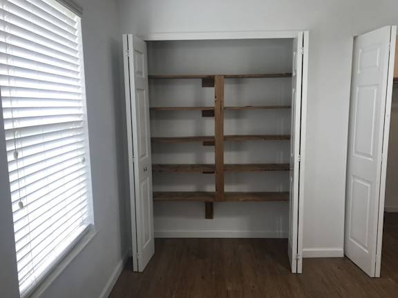 A white closet with a wooden shelf and a window with blinds.