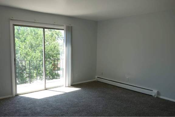 A room with a carpeted floor, a window with a view of trees, and a white wall.