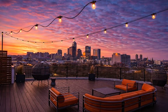 A rooftop patio with orange furniture and string lights overlooking a city skyline at sunset.