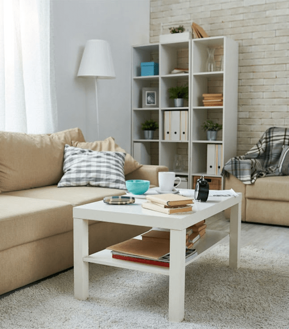 A living room with a beige couch, a white coffee table, and a white shelf with various items on it.