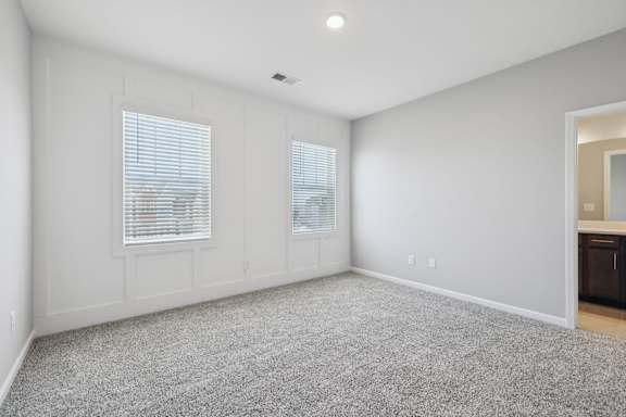 A spacious room with a grey carpet and two windows with blinds.