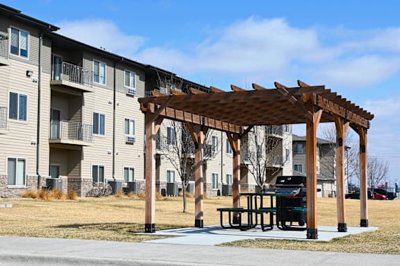 A wooden pergola with a bench is in front of apartment buildings.