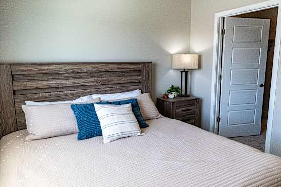 Bedroom with bed, wood headboard, nightstand, table lamp and carpet flooring at Heartland Flats Alliance.
