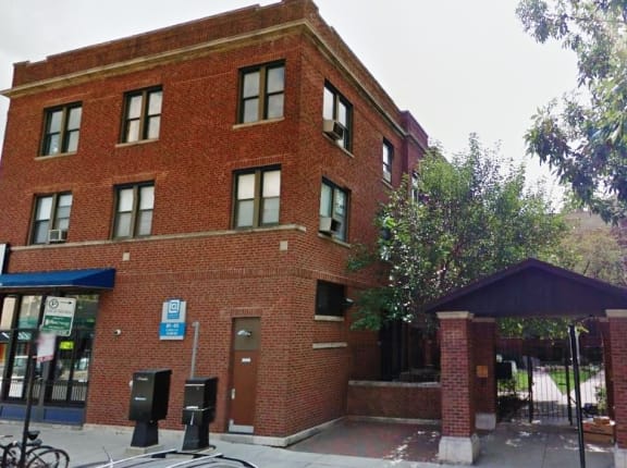Historic three-story red brick building on a corner, with trees, a sidewalk, and a covered side entrance.