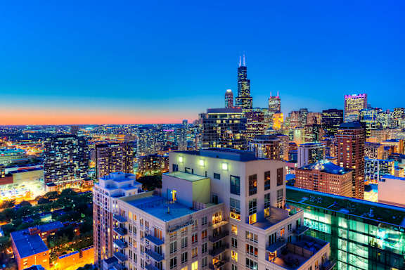 A cityscape at dusk with buildings illuminated against a darkening sky.