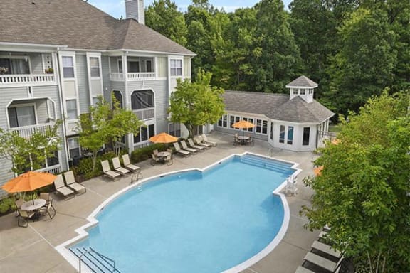 Resort-style outdoor pool area surrounded by lounge chairs and umbrellas, with apartment buildings and mature trees in the background.