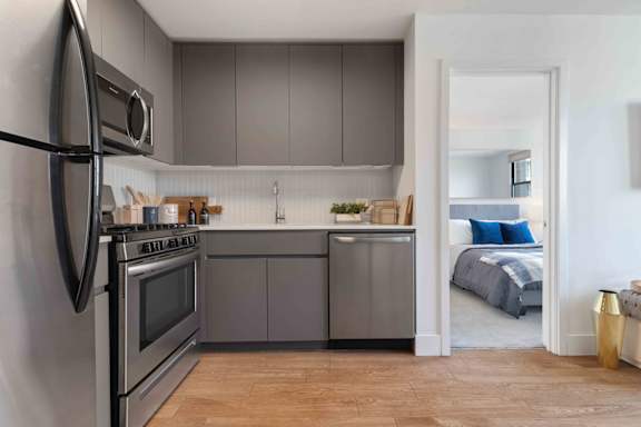 A modern kitchen with a stainless steel refrigerator and oven, and a bedroom visible through the open door.