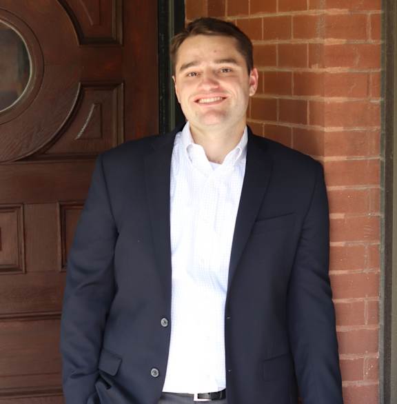 A man in a suit stands in front of a brown door.