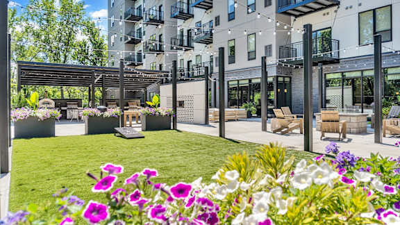 A patio area with a table and chairs is surrounded by a grassy area with purple flowers.