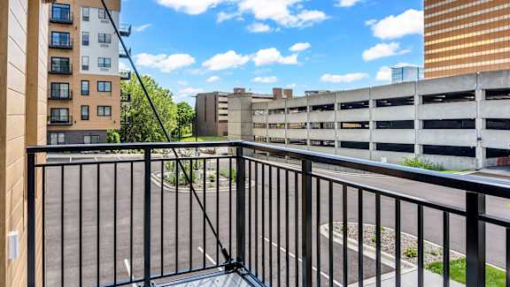 A balcony with a black railing overlooks a parking garage and buildings.