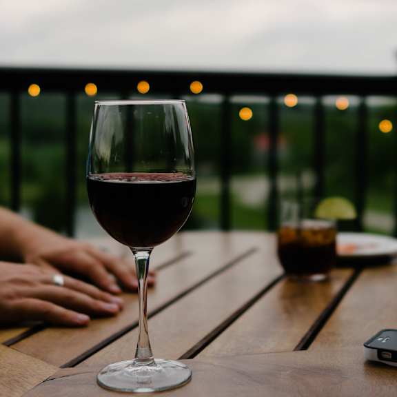 A glass of red wine is on a table with a person's hands in the background.