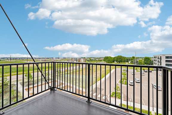 A balcony with a black railing overlooks a parking lot and buildings.