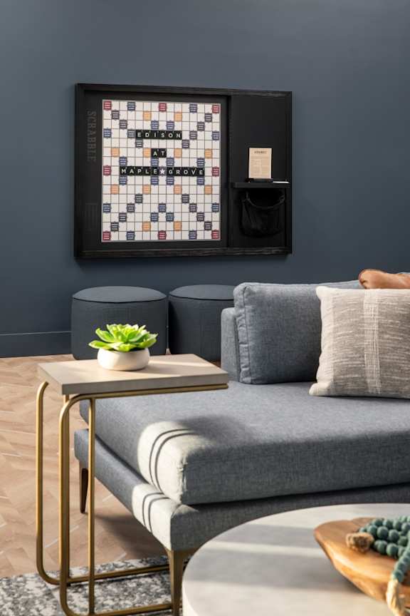 A living room with a grey couch, a small table with a plant and a book, and a framed board game on the wall.at The Edison at Maple Grove, Minnesota
