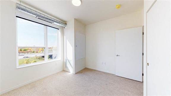A room with a carpeted floor, a window with blinds, and a door at The Bessemer at Seward Commons Apartments, Minneapolis
