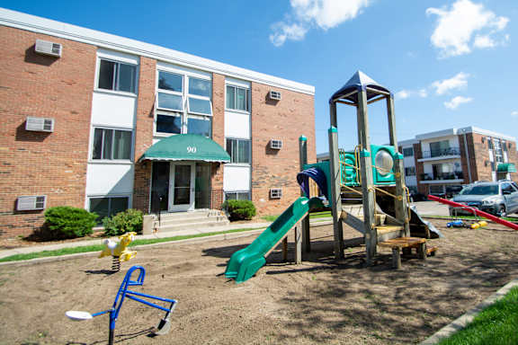 A playground with a green slide and a wooden swing set in front of a brick building with the number 90 on it.