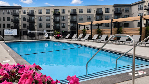 A pool surrounded by chairs and flowers with apartment buildings in the background.