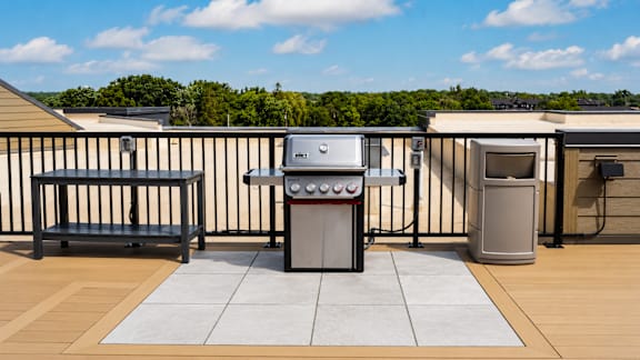 A BBQ grill is on a patio with a bench and a trash can.