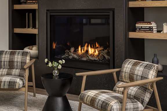 A fireplace with a fire burning inside and two chairs with plaid upholstery in front of it.