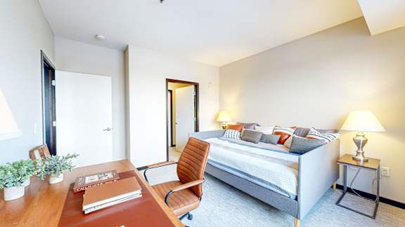 A bedroom with a bed, nightstand, chair, and a desk with a plant and a book on it.