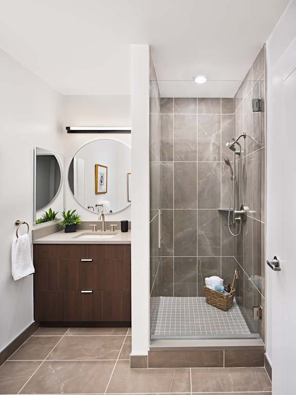 A bathroom with a tiled shower and a round mirror above the sink.