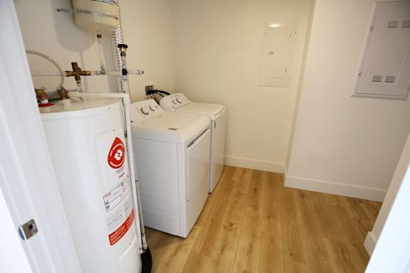 A white fridge with a red and white sticker on it sits in a small room with a washing machine and a white wall.