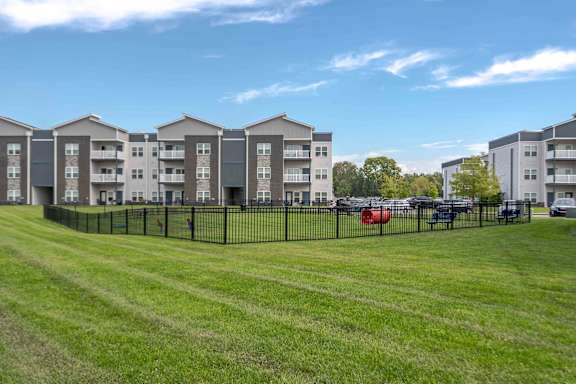 A row of apartment buildings with a grassy field in front and cars parked in the background.