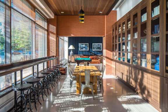 A long wooden table is surrounded by chairs in a room with a brick wall and a long window. at The Maximilian Apartments, Long Island City