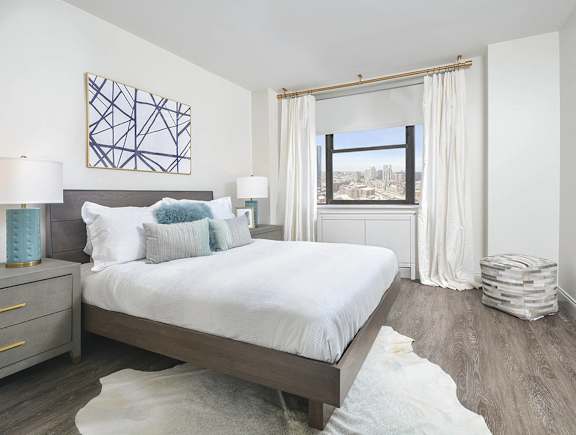 A bedroom with a large bed, a nightstand with a lamp, and a window with a view of the city.
