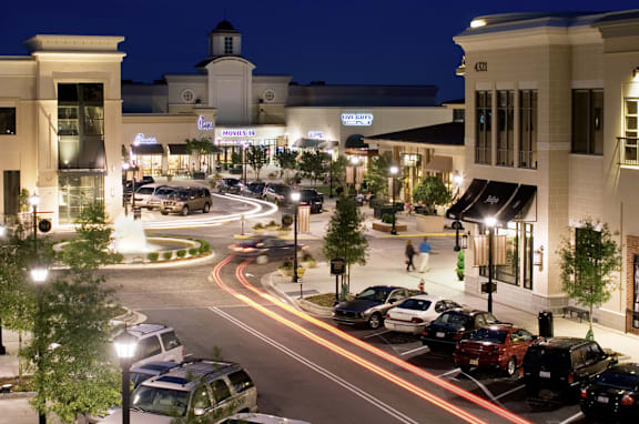 A street view of a shopping area at night with cars parked on the side of the road.