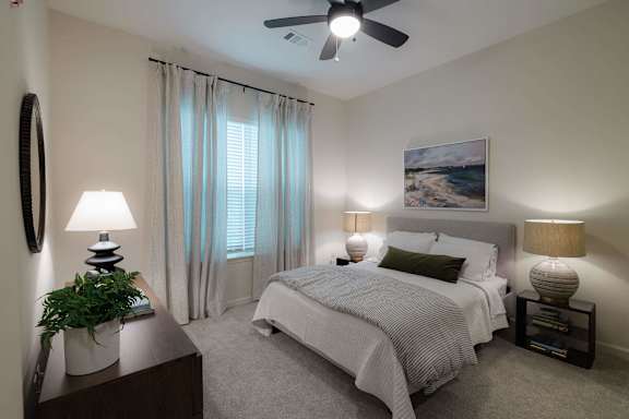 A bedroom with a large bed, a ceiling fan, and a painting on the wall at Hudson at Renaissance Apartments, Wilmington, NC