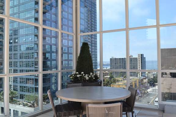 A round table with chairs and a tree in a pot is in front of a large window with a view of the city.