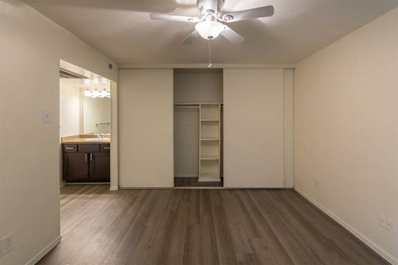 The bedroom with white ceiling fan and a view of teh bedroom closet with built-in shelving in a 1 bedroom, 1 bath C, 653 sq ft apartment home with hardwoodlike flooring throughout at The Biltmore Apartments located in the Vickery Midtown neighborhood of Dallas, TX.