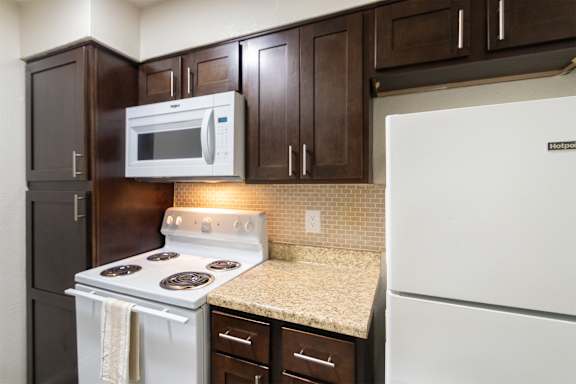 A kitchen with dark wood cabinets and white appliances in a 1 bedroom, 1 bath B, 629 sq ft apartment home at The Biltmore Apartments located in the Vickery Midtown neighborhood of Dallas, TX.