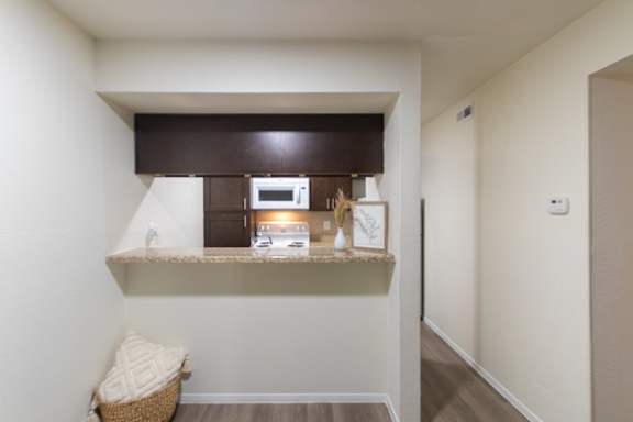 A kitchen with dark wood cabinets and white appliances and breakfast bar viewed from the dining area of a 1 bedroom, 1 bath B, 629 sq ft apartment home at The Biltmore Apartments located in the Vickery Midtown neighborhood of Dallas, TX.