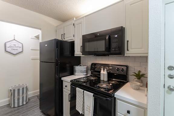 Kitchen view in a 1-bedroom apartment at Cambridge Court Apartments in Dallas, Texas, showing white cabinetry, black appliances, range, microwave, and pantry area.