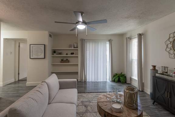 Living room in a 1-bedroom apartment at Cambridge Court Apartments in Dallas, Texas, showing wood-style flooring, ceiling fan, patio door, and built-in shelving.