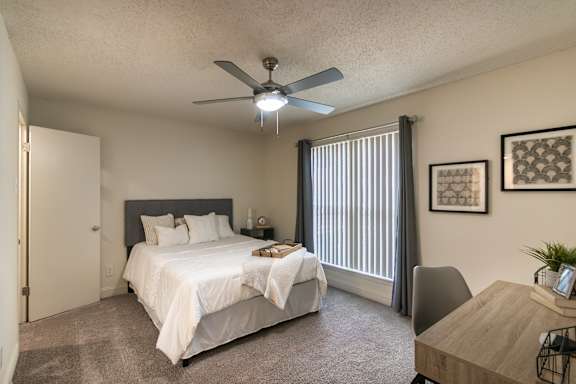 Bedroom in a 1-bedroom apartment at Cambridge Court Apartments in Dallas, Texas, showing carpeted flooring, ceiling fan, patio door, and staged furniture.