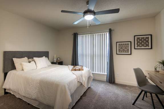 Staged bedroom in a 1-bedroom apartment at Cambridge Court Apartments in Dallas, Texas, featuring carpeted flooring, ceiling fan, patio door, desk area, and natural light.