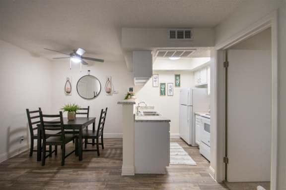 A kitchen with a white cabinets and appliances and the dining room with table and chairs in a 515 square foot 1 bedroom, 1 bath apartment home at Canyon Creek Apartments in Dallas Midtown neighborhood of Dallas, TX.