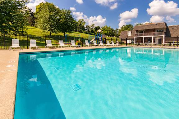 A pool surrounded by white lounge chairs with the playground in the background at Fairfield Pointe Apartments in Fairfield, Ohio.