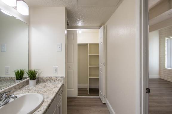 A hallway closet with a view of white bath vanity and a look into the bedroom with hardwoodlike flooring in a 1 bedroom, 1 bath apartment home at Harvard Square Apartments in the Vickery Midtown neighborhood of Dallas, TX.