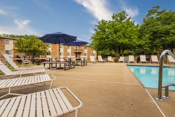 A pool area with sun loungers, picnic tables with umbrellas at Blue Grass Manor Apartments in Erlanger, KY.