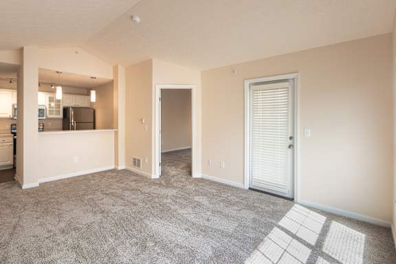 Open living room at Nantucket Apartments with carpeted flooring, patio door, kitchen breakfast bar, and natural light across the room.