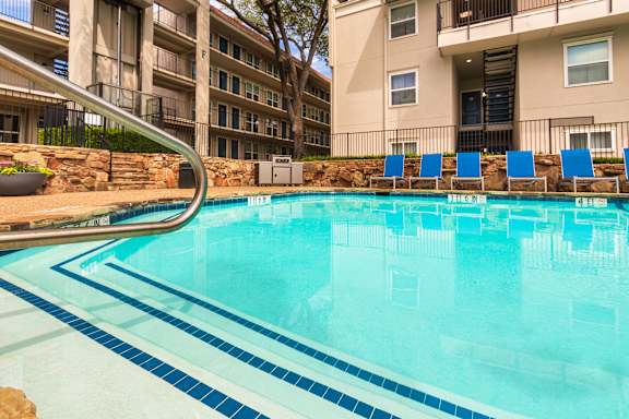A pool area with lounge chairs, a patio table and chairs with a blue umbrella and stainless steel BBQ in front of apartment buildings at Princeton Court Apartments in the Vickery Midtown neighborhood of Dallas, TX.