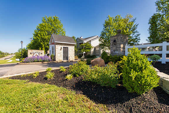 A small stone building with a black door is surrounded by a garden with a white fence in the background.