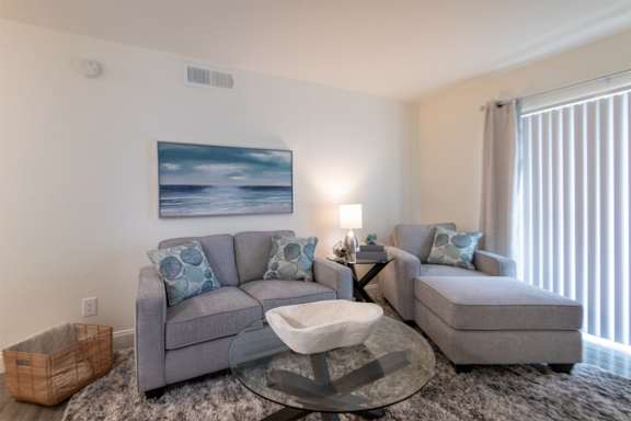A modern living room with a grey sofa, chaise lounge and a glass coffee table in the 558 square foot 1 bedroom, 1 bath model apartment at The Summit at Midtown Apartments in the Dallas Midtown neighborhood of Dallas, TX.