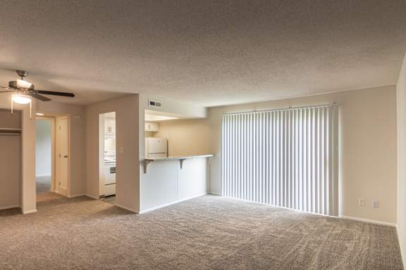 Wide living room view at Village East Apartments in Franklin, Ohio, with vertical blinds, carpeted flooring, kitchen pass-through, and open apartment layout.