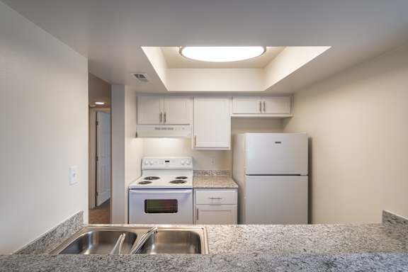 Kitchen counter view at Village East Apartments, looking across the double sink toward white cabinets, refrigerator, and electric range.