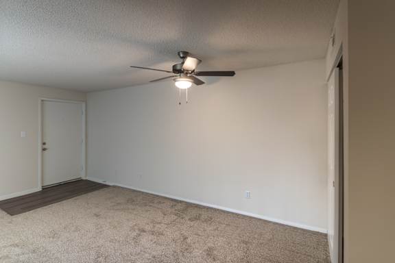 Living area in the Village East 1-bedroom apartment, with carpeted flooring, ceiling fan, neutral walls, and kitchen nearby.